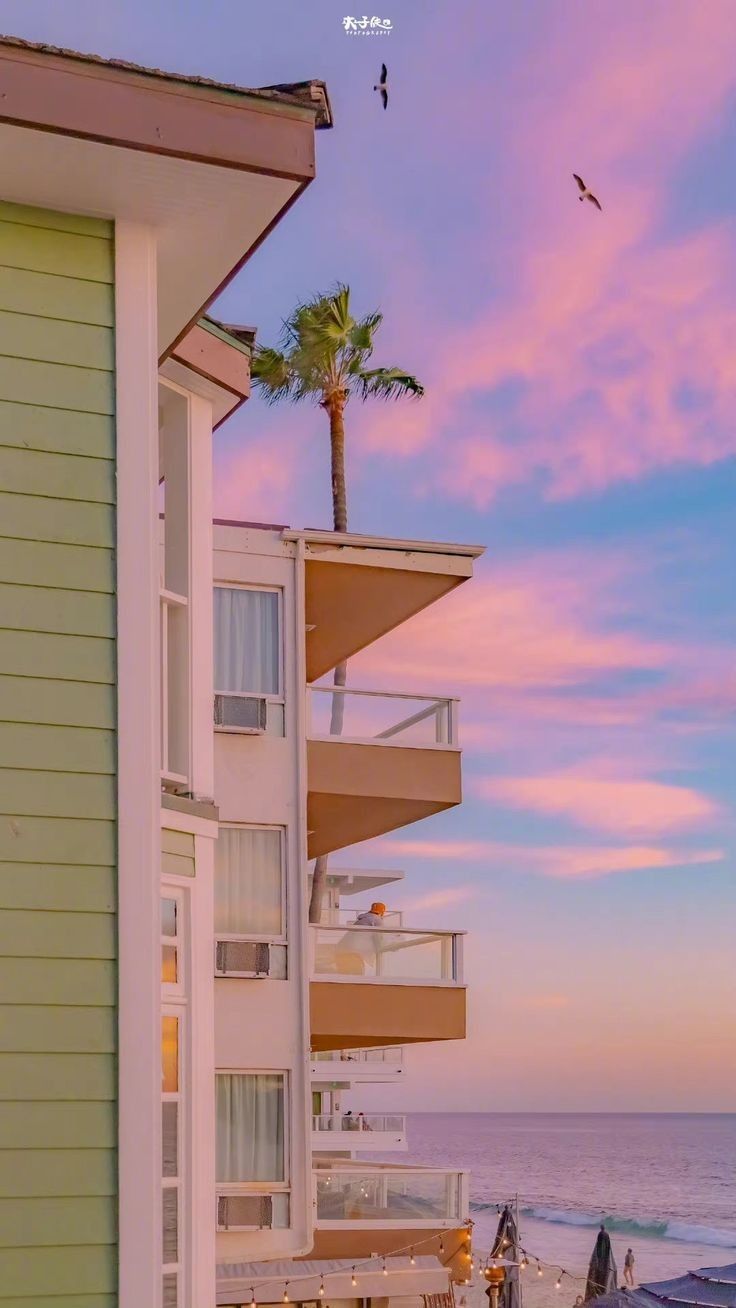 A pastel apartment building with balconies and a palm tree under a pink sky by the sea.