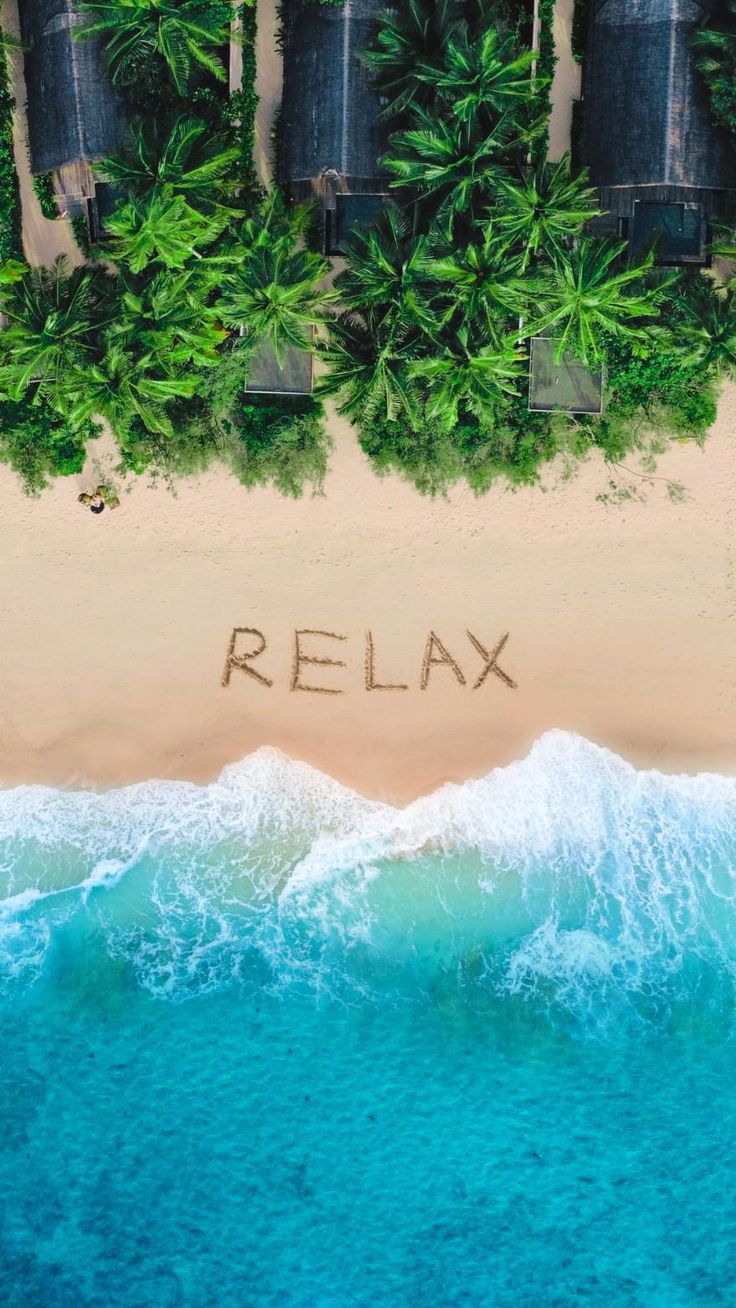 "Relax" written in the sand above turquoise waves and green trees, seen from above.