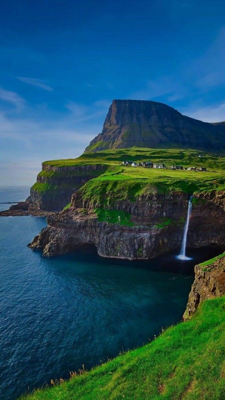 A waterfall pouring off a green cliff into the sea beneath a tall mountain.