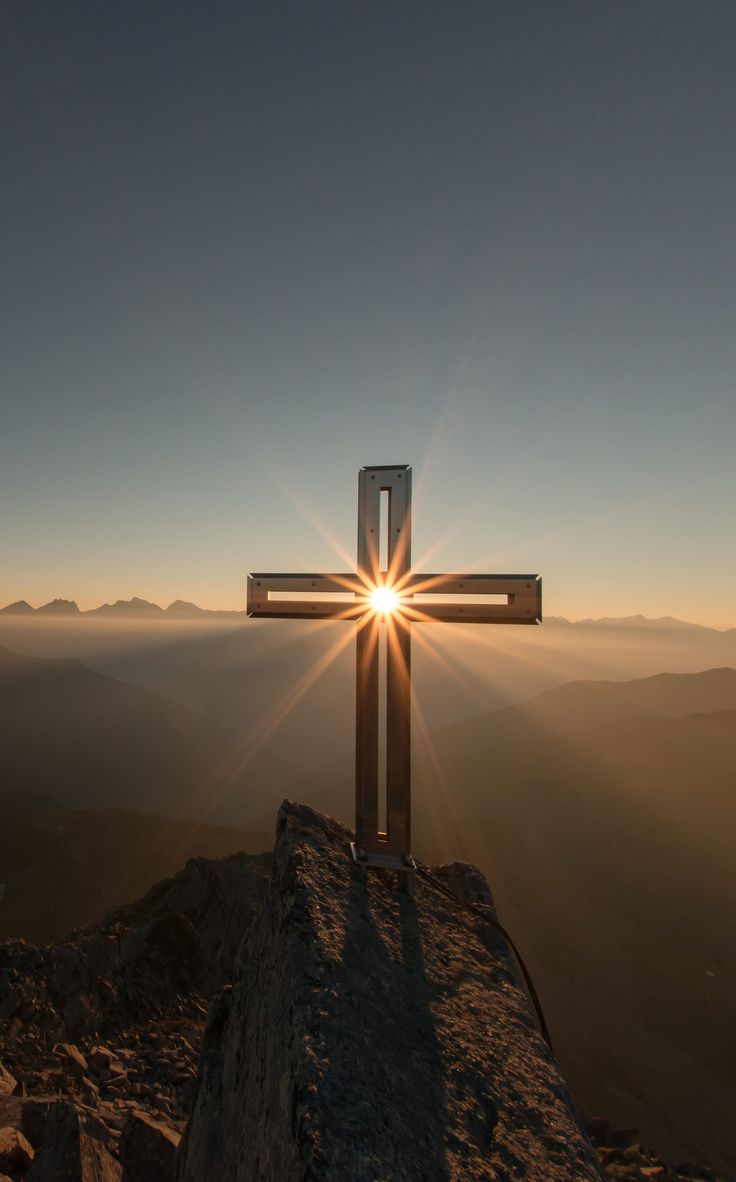 A wooden cross on a rocky peak with the sun bursting behind it.