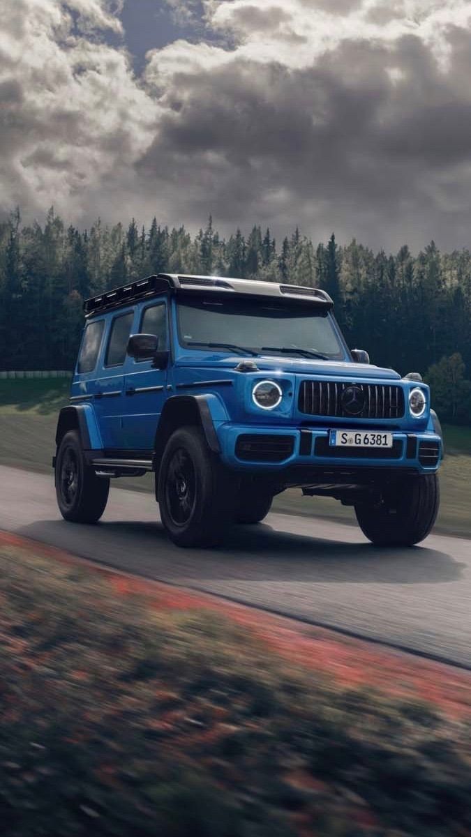 A blue Mercedes G-Class on a road beside pine trees under grey clouds.