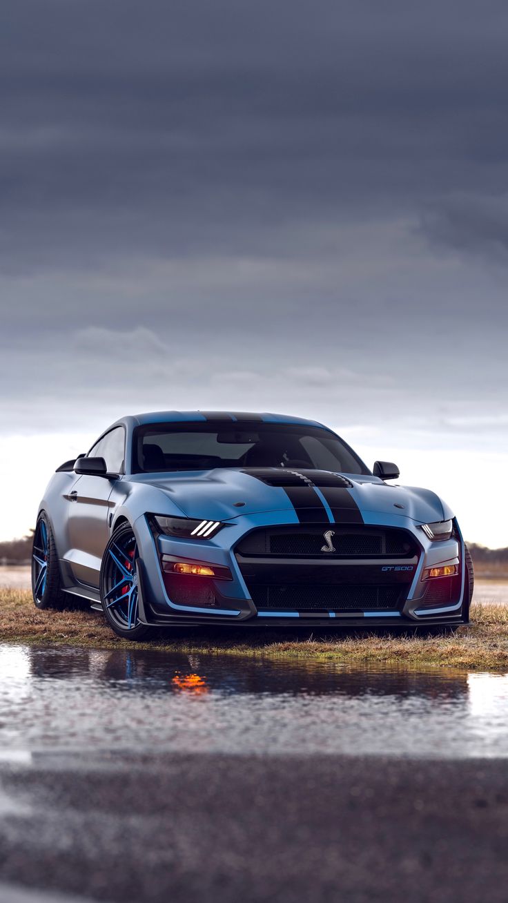 A blue Ford Mustang Shelby GT500 on a wet road under a stormy sky.