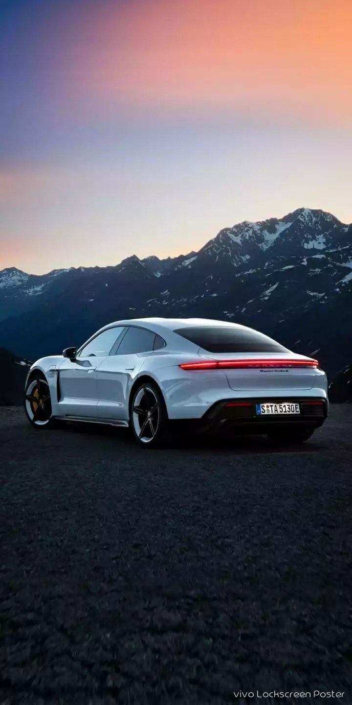 A white Porsche Taycan parked on dark asphalt with mountains behind at dusk.