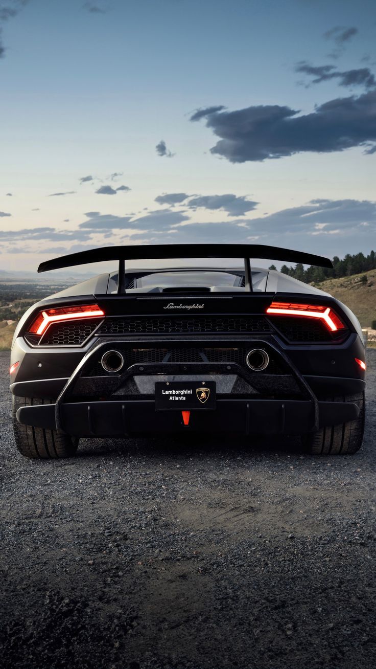 A black Lamborghini Huracán seen from behind with its big rear wing, under a cloudy evening sky.