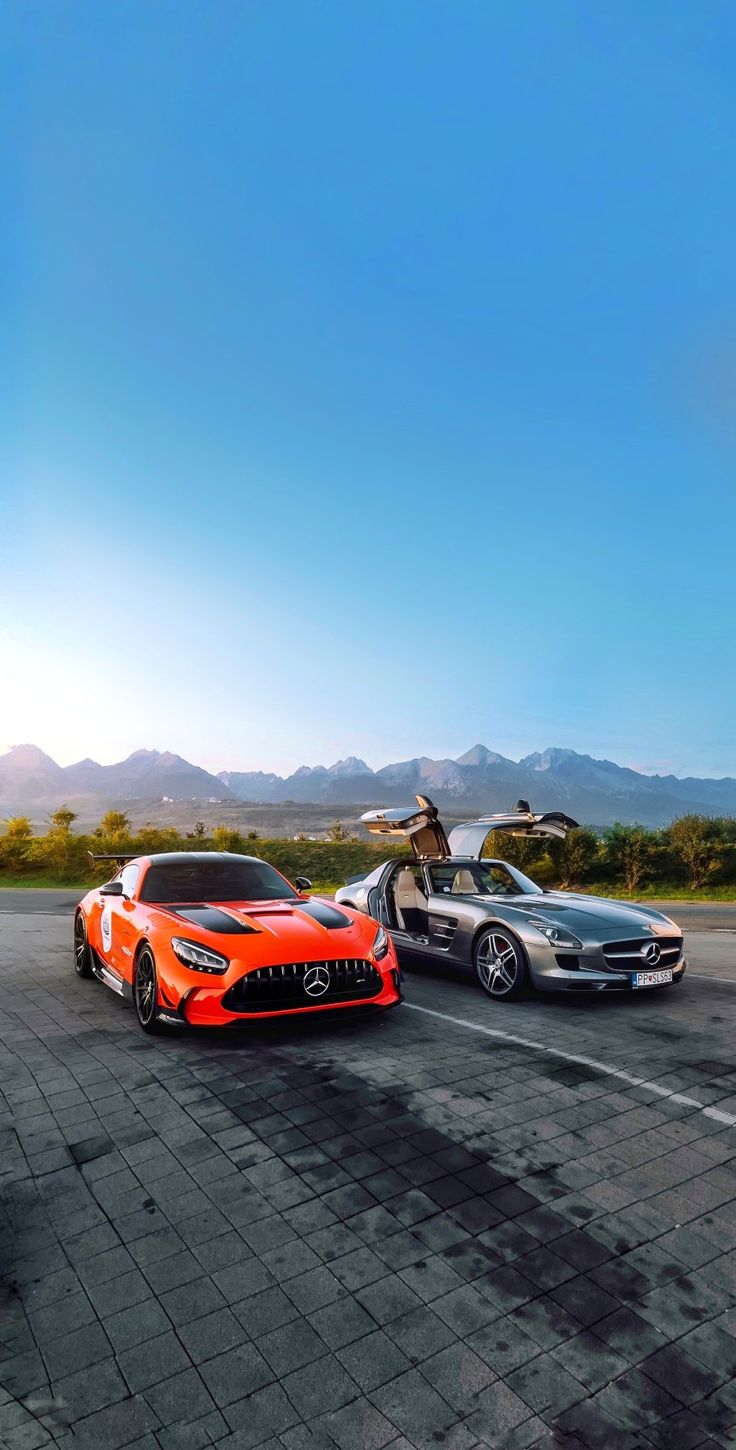 An orange and a silver Mercedes-AMG parked on a wet road with mountains behind.