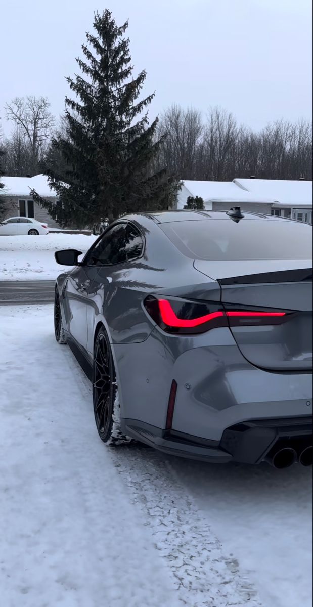A grey BMW M4 parked on a snowy driveway beside pine trees.